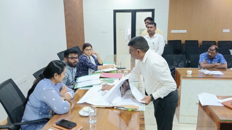 ಬಿಳಿ ಅಂಗಿ ಧರಿಸಿದ ವ್ಯಕ್ತಿಯೊಬ್ಬರು ಸಭಾಂಗಣದಲ್ಲಿ ಮೇಜಿನ ಮೇಲಿನ ಕಾಗದಗಳನ್ನು ಪರಿಶೀಲಿಸುತ್ತಿದ್ದು, ಸುತ್ತ ಹಲವರು ಕುಳಿತಿದ್ದಾರೆ.